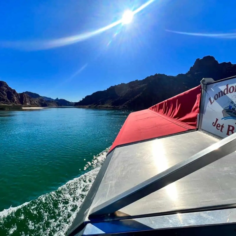 Boat cruising on a river with rocky hills on a sunny day, sign reads 'London Bridge Jet Boat'.