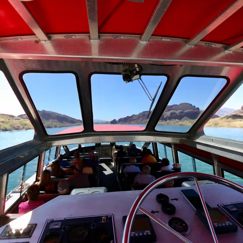 View from inside a boat with passengers, overlooking a lake and mountains through large windows.