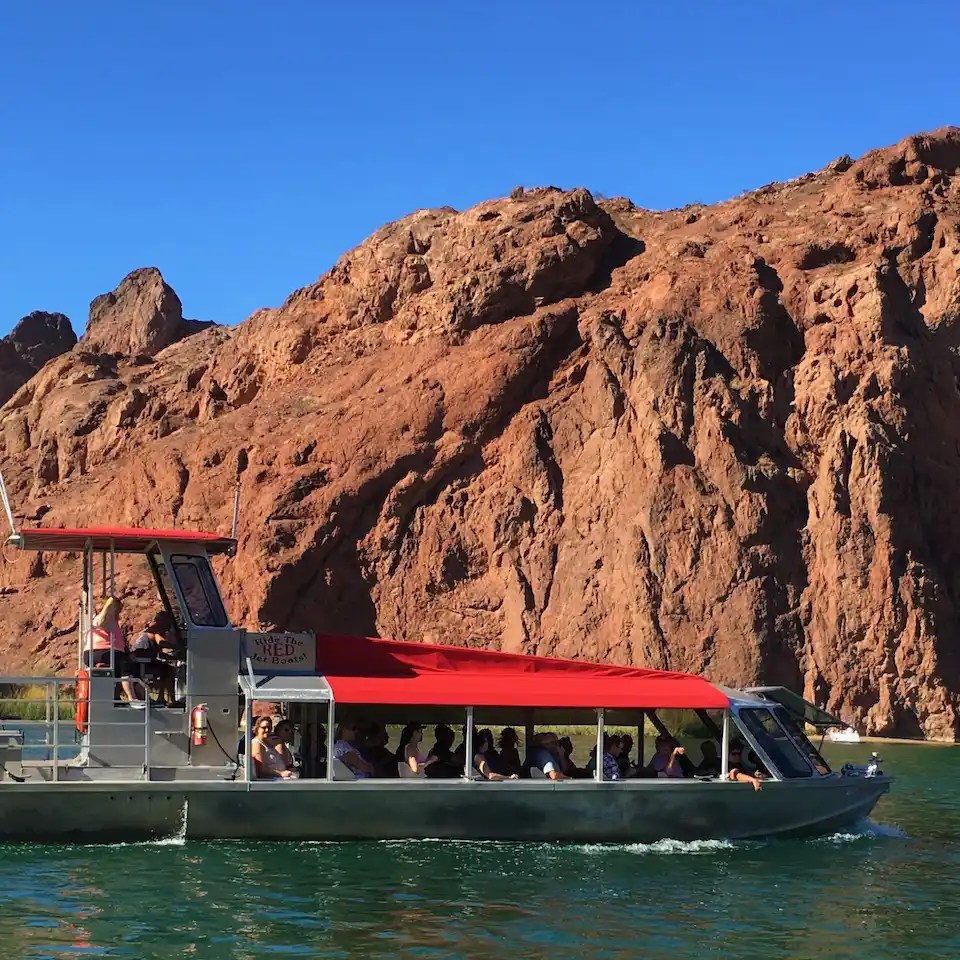 Tour boat with red canopy on a river near rocky cliffs under blue sky.