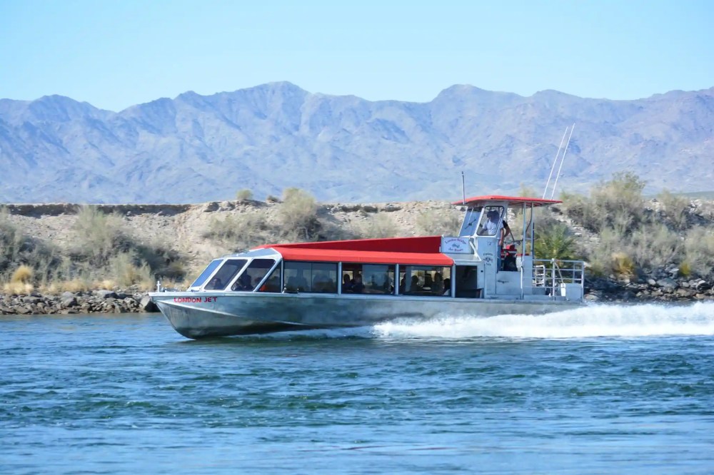 Jet boat with red canopy cruising on a river, mountains in the background.