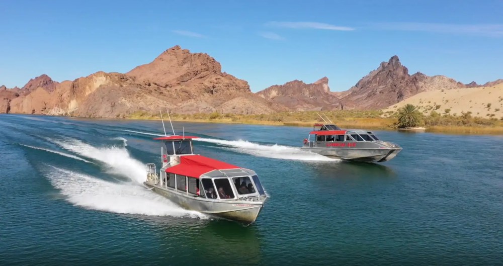 Two speedboats with red canopies racing on a river near rocky desert landscape.