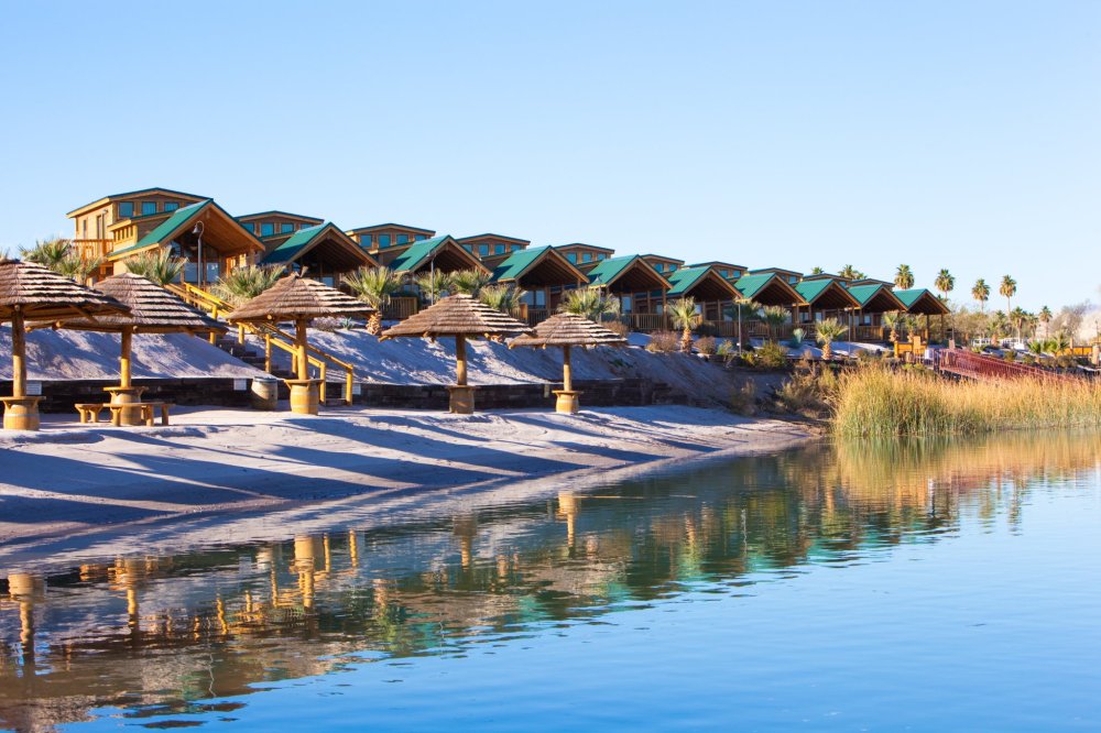 Rows of lakeside cabins with shaded picnic areas and palm trees by the water under a clear blue sky.