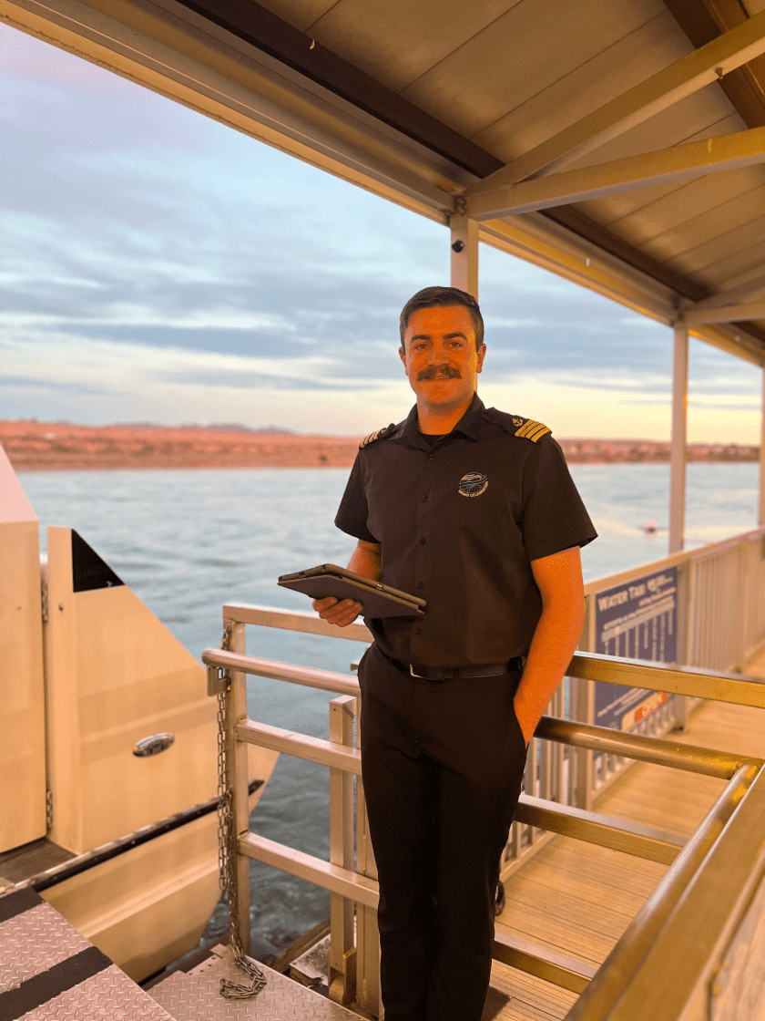 Uniformed man standing on a dock by the water at sunset, holding a tablet.